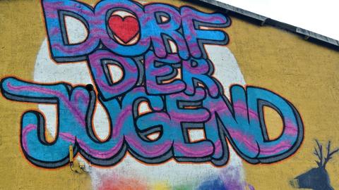 Schriftzug "Dorf der Jugend" in Blau und Pink an eine Mauer gesprayt, im "o" ein rotes Herz, im Hintergrund eine Wolke, aus der ein Regenbogen kommt, sowie ein Hirschkopf