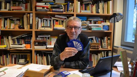 Prof. Dr. Peter Gendolla sitzt vor einer Bücherwand am Schreibtisch und hält Karten in den Händen.