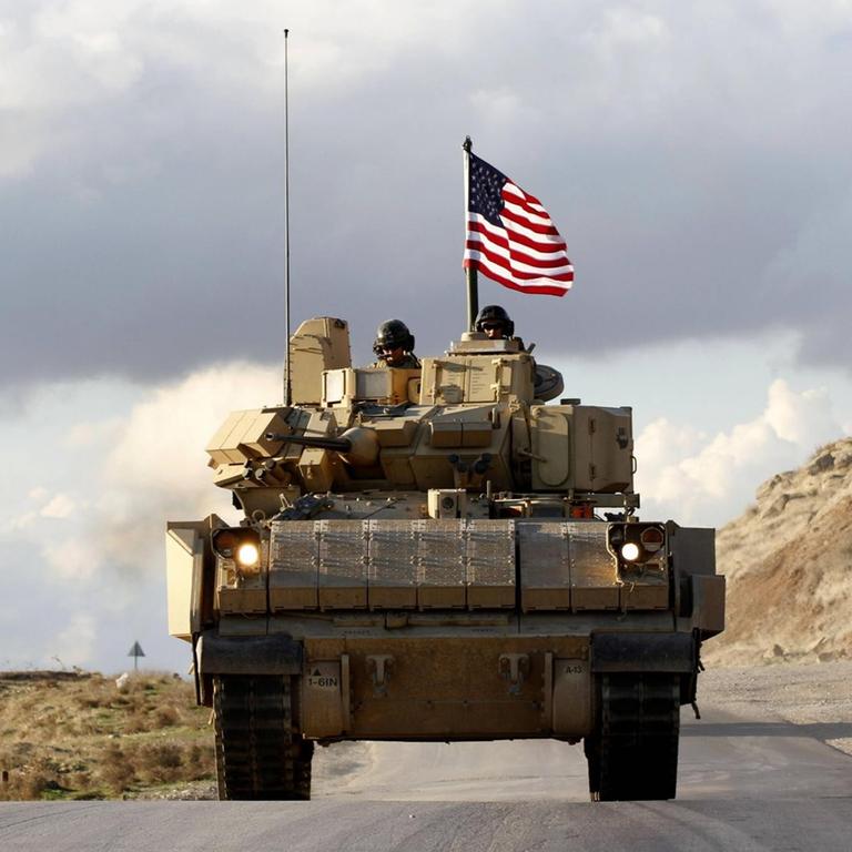 Ein Patrouillenfahrzeug der Joint Task Force CJTF-OIR mit US-Soldaten fährt auf einer Wüstenstraße. Oben auf dem Wagen weht eine US-Flagge.