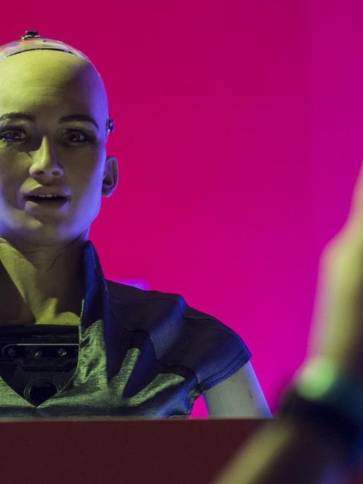 "Sophia the Robot" steht hinter einem Podium, mit einer pinken Fläche im Hintergrund und wird mit einem Smarphone fotografiert.