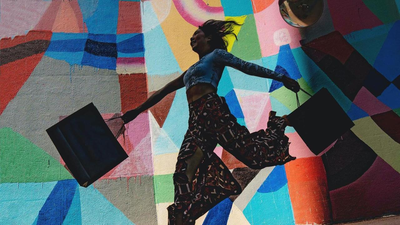 Eine Frau springt mit ihren Einkaufstüten tänzerisch vor einer bunten Fassade.