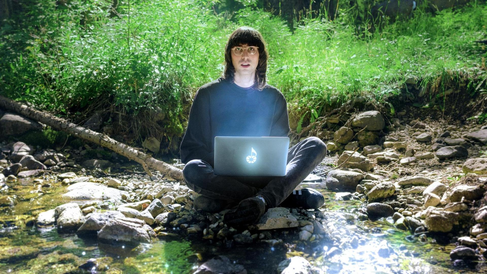 A.G. Cook sitzt mit einem geheimnisvoll leuchtenden Laptop auf dem Schoß im Schneidersitz in einem Waldflussidyll und blickt erstaunt in die Kamera.