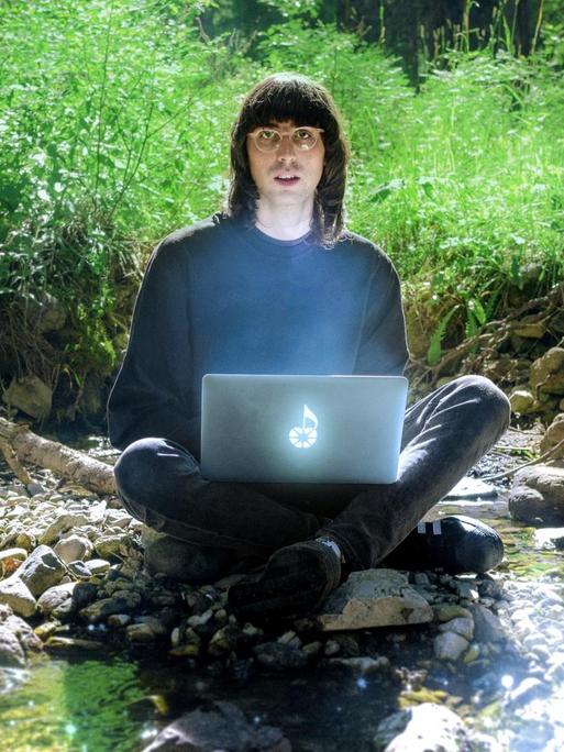 A.G. Cook sitzt mit einem geheimnisvoll leuchtenden Laptop auf dem Schoß im Schneidersitz in einem Waldflussidyll und blickt erstaunt in die Kamera.