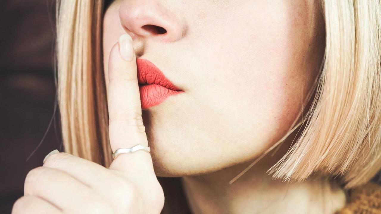 Eine blonde junge Frau legt ihren Zeigefinger auf die Lippen und symbolisiert Schweigen.