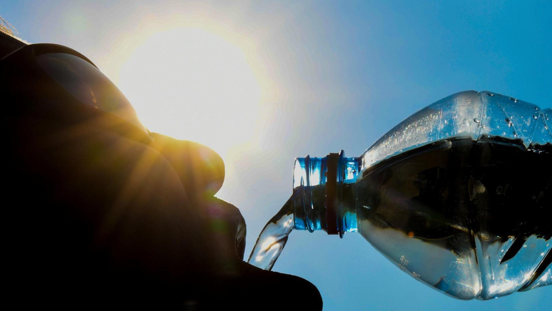 Ein Mann lässt Wasser aus einer Flasche in seinen Mund laufen. Das Foto ist gegen die Sonne und vor blauem Himmel aufgenommen worden.