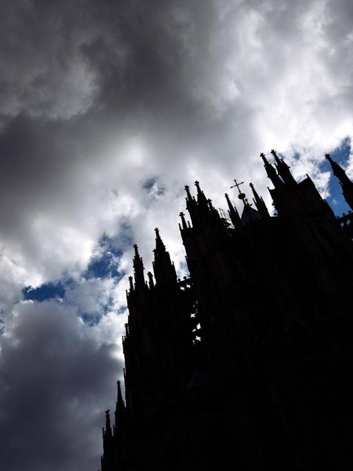 Sturmwolken ziehen auf über dem Kölner Dom. Das Gebäude ist aus der Froschperspektive nur als Schattenriss erkennbar, darüber brauen sich dunkle Wolken zusammen.