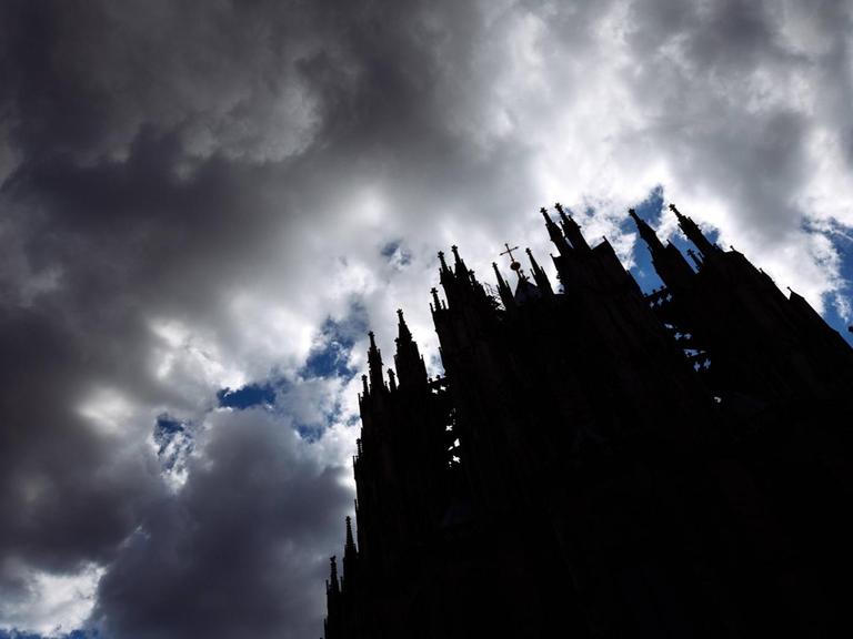 Sturmwolken ziehen auf über dem Kölner Dom. Das Gebäude ist aus der Froschperspektive nur als Schattenriss erkennbar, darüber brauen sich dunkle Wolken zusammen.