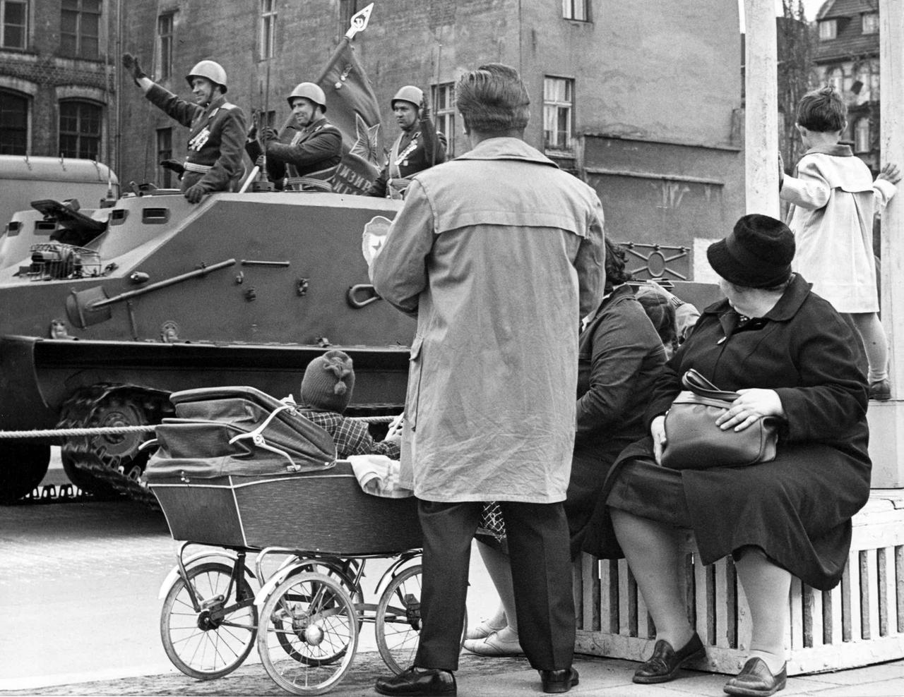 Das Schwarz-Weiß-Foto zeigt Zzuschauer einer Parade, im Hintergrund Soldaten in Uniform auf einem vorbeifahrenden Panzer.