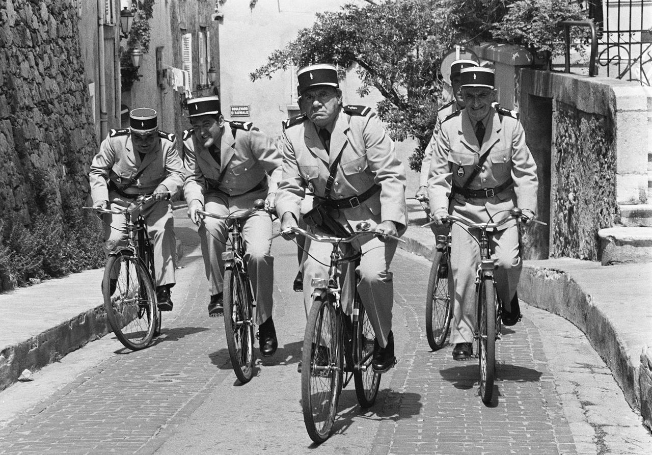Man sieht den Schauspieler Michel Galabru (Mitte) an der Seite von Louis de Funès (rechts) auf dem Fahrrad - im Film "Louis und seine verrückten Politessen".