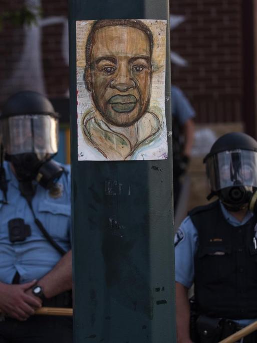 Ein Porträt von George Floyd hängt an einem Straßenlaternenmast, während Polizeibeamte bei einer Gegenüberstellung mit einer Gruppe von Demonstranten in Minneapolis, Minnesota, Wache stehen.