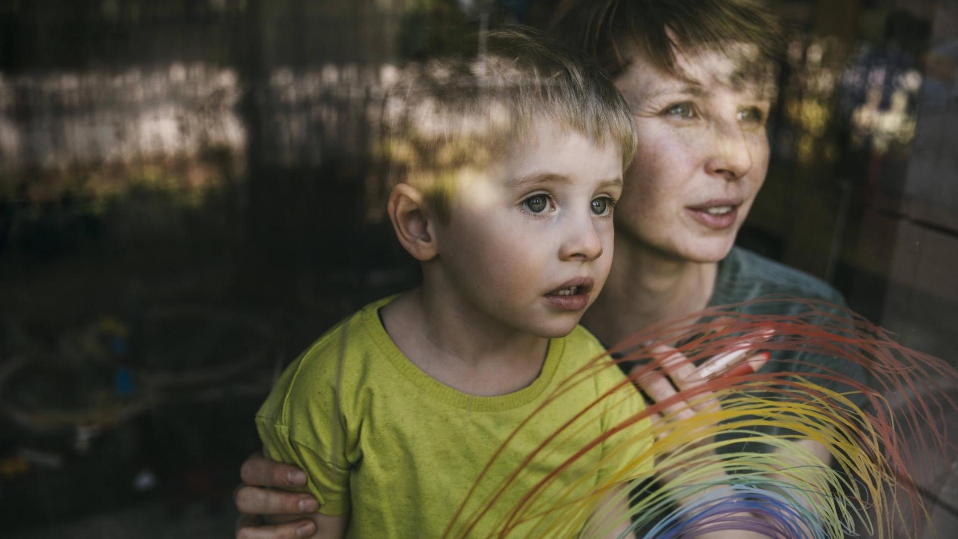 Eine Frau hält einen kleinen Jungen von hinten an den Schultern – beiden schauen zum Fenster hinaus, an das mit dünnen Strichen ein bunter regenbogen gemalt ist.