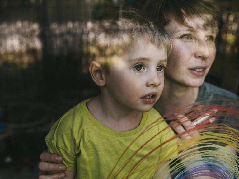Eine Frau hält einen kleinen Jungen von hinten an den Schultern – beiden schauen zum Fenster hinaus, an das mit dünnen Strichen ein bunter regenbogen gemalt ist.