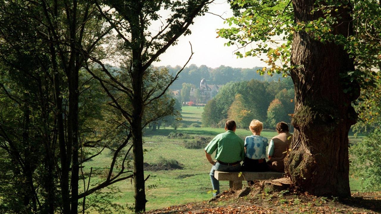 Auf der polnischen Seite des Pücklerparks in Leknica sitzen Spaziergänger auf der Steinbank am Mausoleumshügel und genießen den Blick entlang der längsten Sichtachse des Parks bis zum Schloss in Bad Muskau auf der sächsichen Seite.