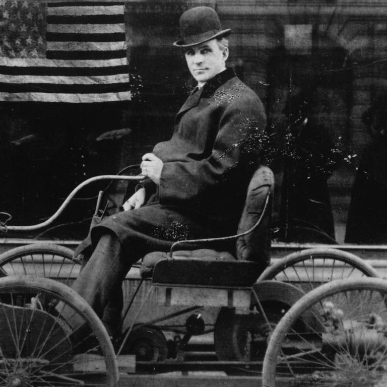 Ein Schwarz-Weiß-Foto zeigt Henry Ford, wie er sein erstes Auto fährt. Im Hintergrund: eine amerikanische Flagge. Henr