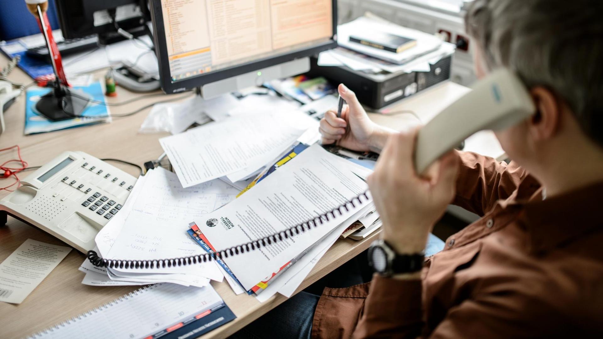 Ein Angestellter sitzt in einem Büro an seinem Schreibtisch und telefoniert vor einem großen Stapel von Unterlagen, aufgenommen am 04.02.2015 in Dresden (Sachsen).