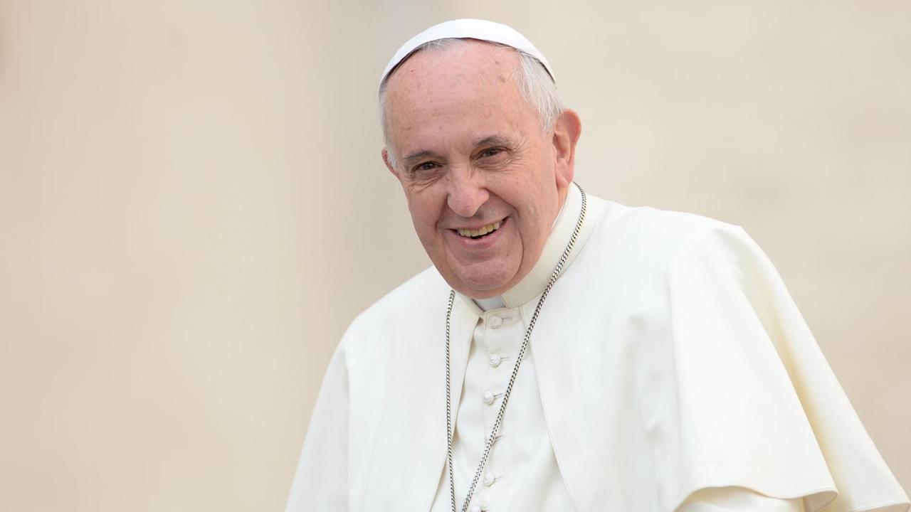 Papst Franziskus am 22.10.2014 auf dem Petersplatz in Rom.
Der Papst lächelt im weißen Gewand und mit umgehängter Kreuzkette vor einer weißen Wand in die Kamera.