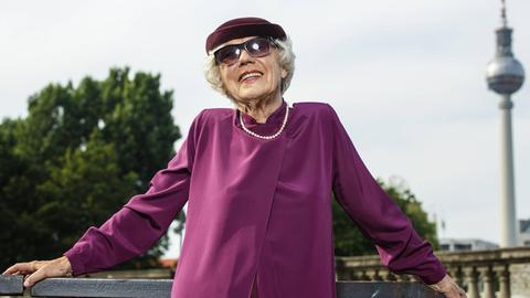 Eine modisch in Violett gekleidete Seniorin mit Perlenkette, Sonnenbrille und Hut posiert vor dem Berliner Fernsehturm