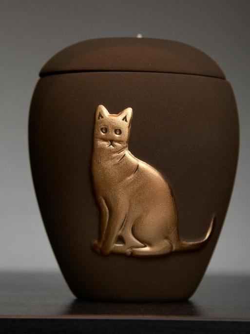 Eine Urne mit dem Relief einer Katze steht in einem Regal.