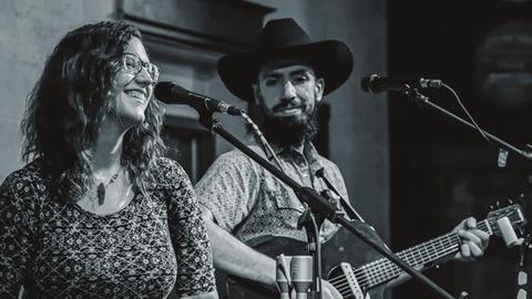 Eine Frau im geblümten Kleid und Brille steht vor einem Mikrofon, neben ihr ein Mann mit Hut, Vollbart und Gitarre in der Hand.