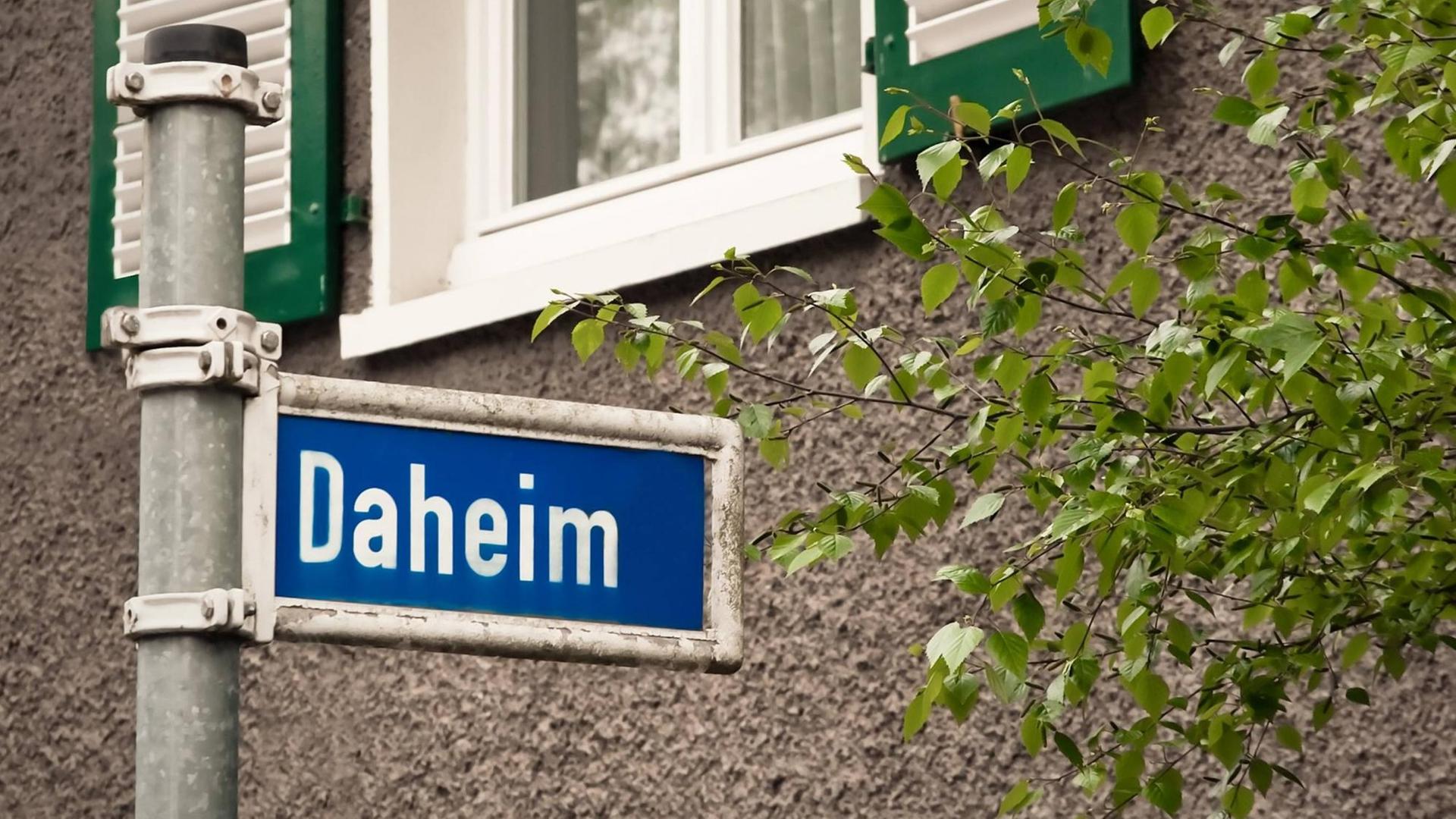 Ein blaues Straßenschild auf dem in weißen Buchstaben "Daheim" steht.