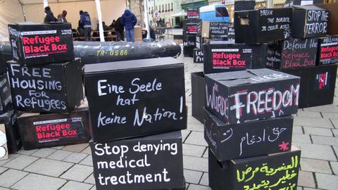 Aufgestapelte schwarze Boxen mit Aufschriften (Refugee Black Box, Eine Seele hat keine Nation!, Stop denying medical treatment, Freedom u.a.), im Hintergrund ein Schlauchboot und mehrere Personen.