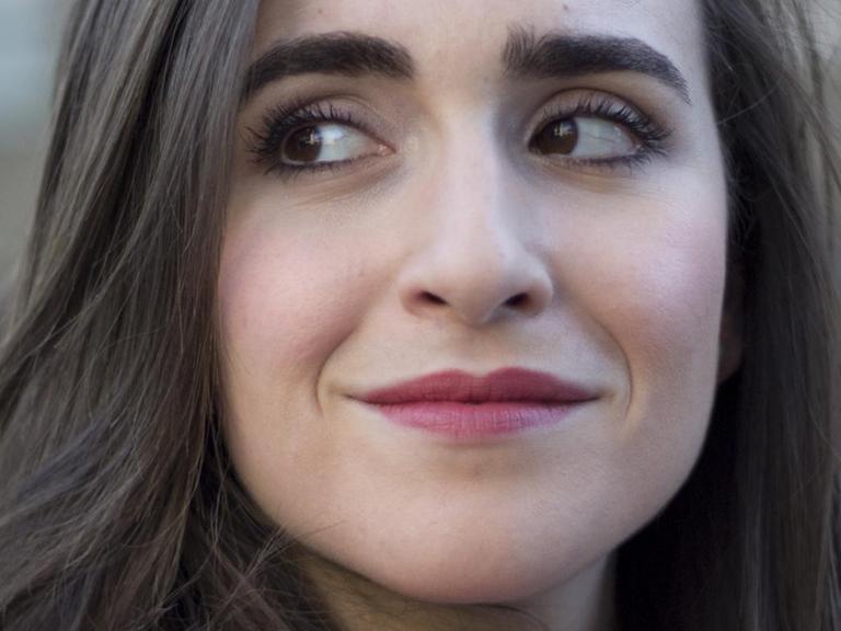 Eine junge Frau mit schulterlangen, braunen Haaren und dunkelrot geschminkten Lippen schaut lächelnd nach links.