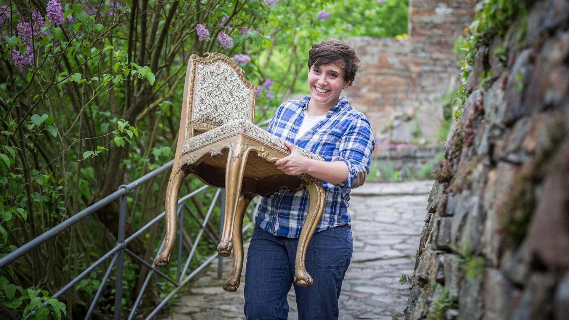 Burgenbloggerin Jessica Schober beim Einzug auf die Burg, Frau trägt einen mondänen Stuhl einen gepflasterten Weg entlang. Burgenbloggerin Jessica Schober beim Einzug auf die Burg, Frau trägt einen mondänen Stuhl einen gepflasterten Weg entlang.