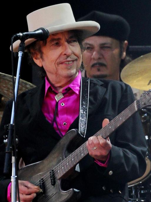 Der Sänger Bob Dylan bei einem Auftritt. Er trägt einen hellen Hut und ein pinkfarbenes Hemd.