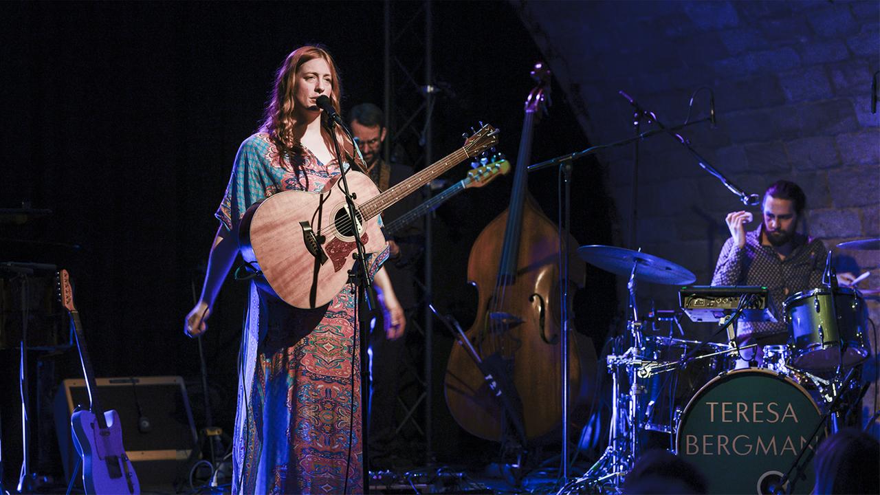 Eine Frau mit roten Locken steht in buntem Kleid auf der Bühne, eine Akustikgitarre vor sich. Hinter ihr Bassist und Schlagzeuger, sowie Kontrabass, E-Gitarre und Verstärker.