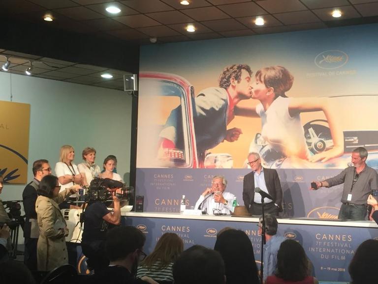 Foto von der Pressekonferenz mit Jean-Luc Godard, der per FaceTime zugeschalten war. Im Hintergrund ist das diesjährige Plakat der Filmfestspiele Cannes zu sehen, das eine Szene aus einem Godard-Film zeigt.