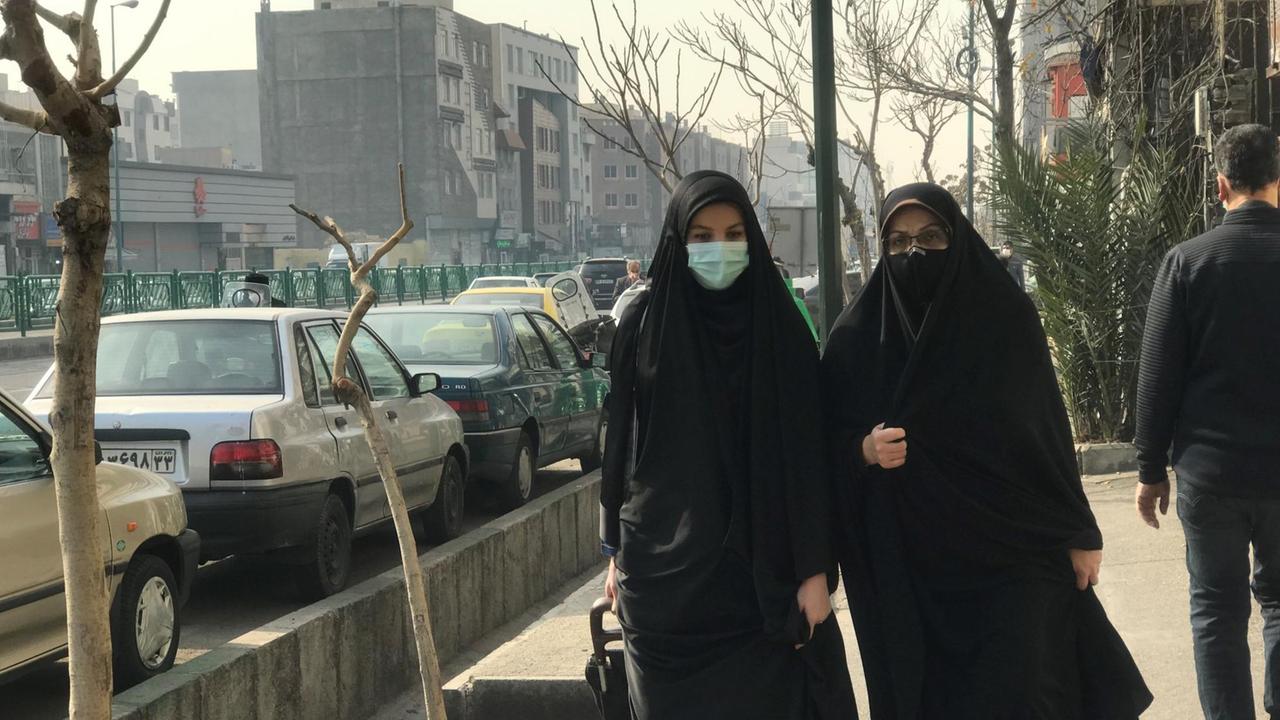 Zwei komplett in schwarzen langen Gewändern und mit Masken verhüllte Frauen gehen mit Einkaufswagen die Straße entlang.