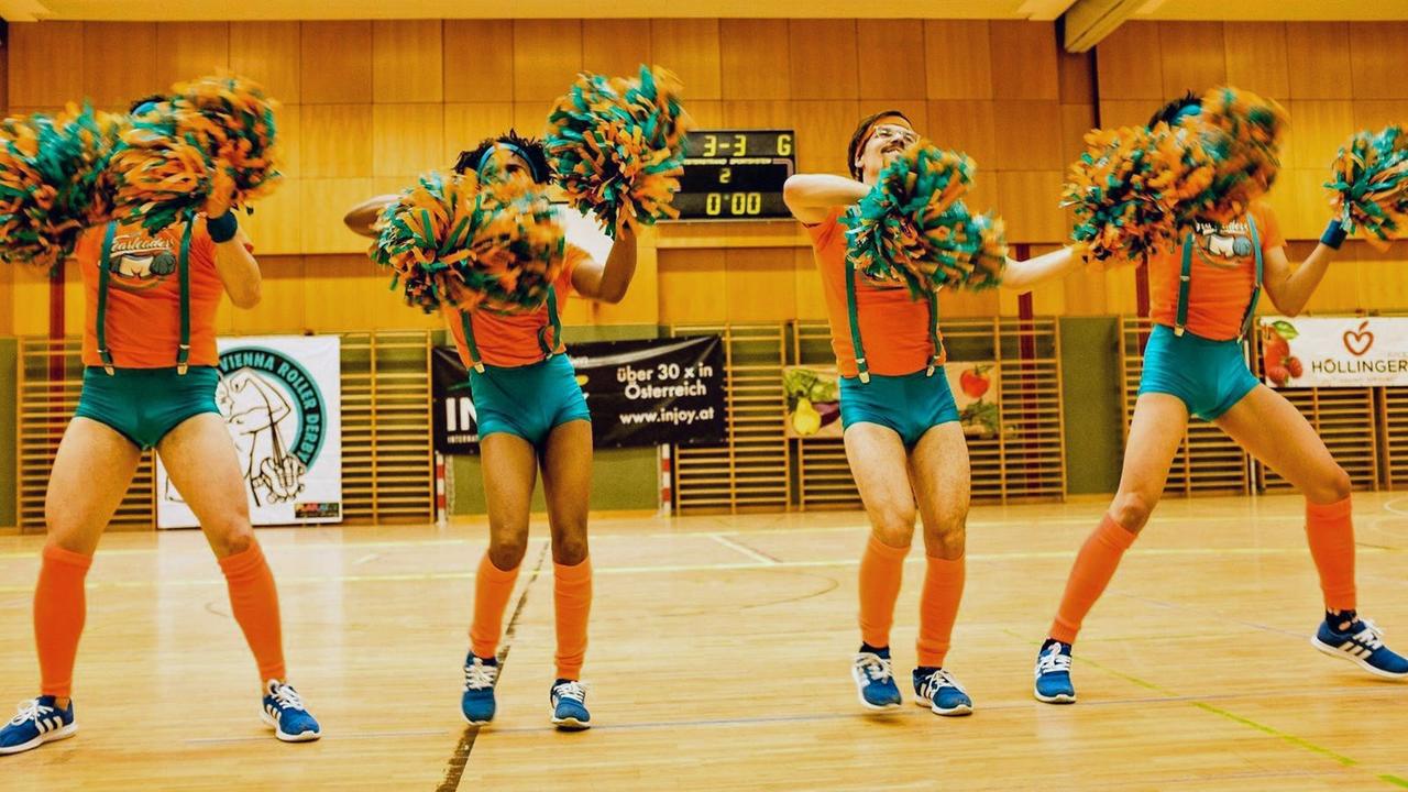Vier Mitglieder der Vienna Fearleaders tanzen in engen türkisen Höschen und mit Pompons in einer Sporthalle.