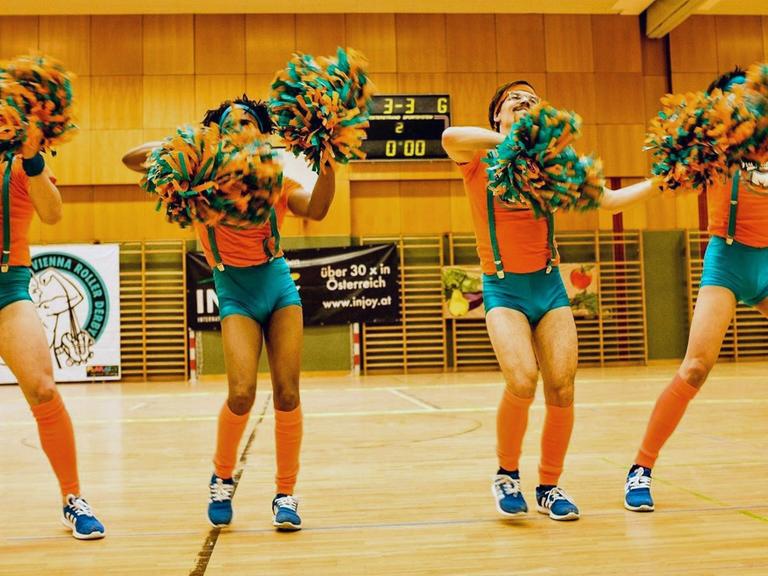 Vier Mitglieder der Vienna Fearleaders tanzen in engen türkisen Höschen und mit Pompons in einer Sporthalle.