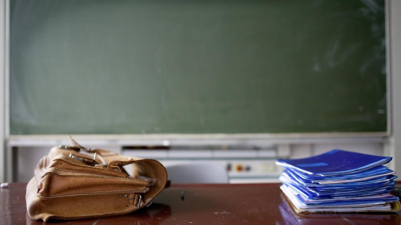 In einem Klassenzimmer liegen eine Aktentasche und ein Heftestapel auf dem Lehrerpult