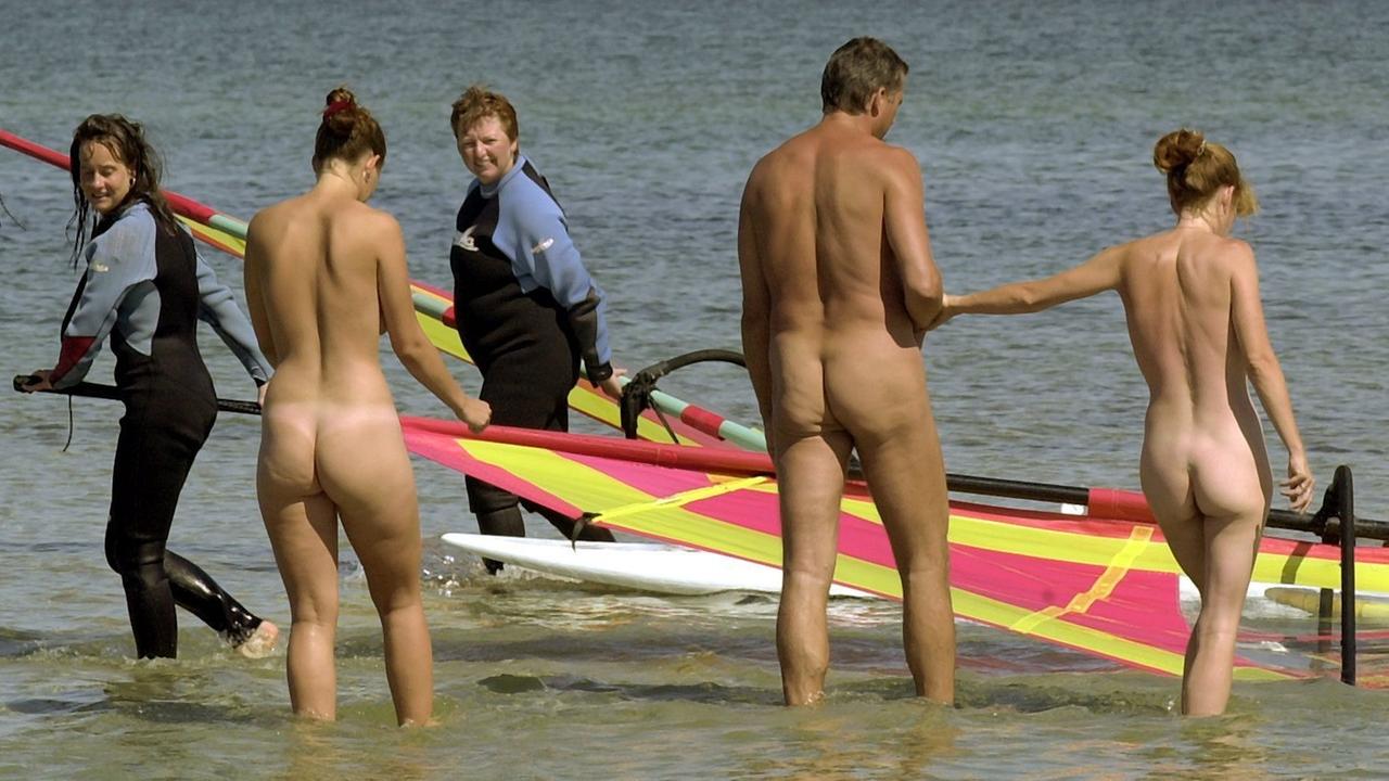 Nackte Schwimmer gehen am Ostseestrand ins Wasser. Zwei Surfer schauen ihnen zu.