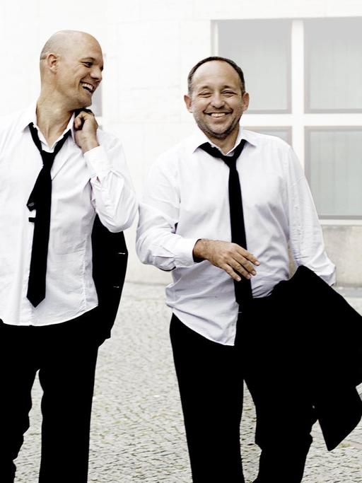 Zwei Männer in weißem Hemd und loser schwarzer Krawatte gehen lachend an einem Gebäude vorbei.
