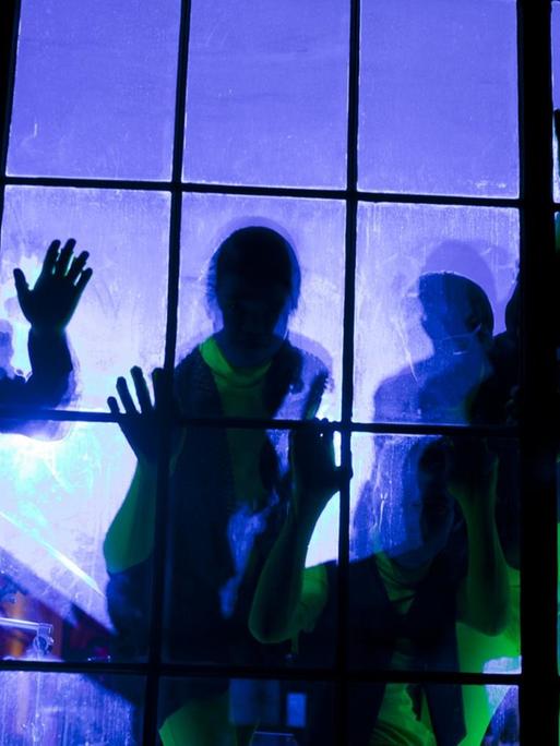 Mehrere Menschen in grünen Hosen stehen hinter einer Fensterscheibe, die Handflächen liegen auf der Scheibe, hinter ihnen lila Licht.