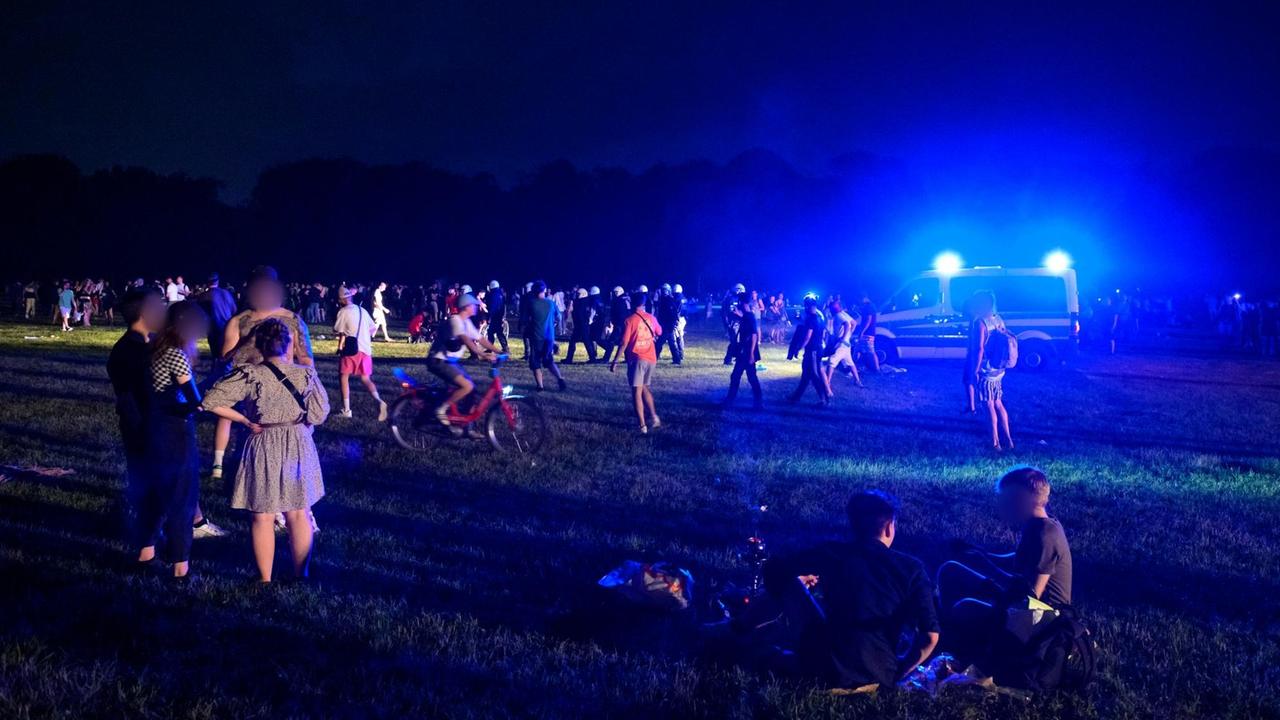 Behelmte Polizisten laufen in Hamburg begleitet von einem Mannschaftswagen mit Blaulicht durch die Menschengruppen auf der Stadtpark-Wiese. 