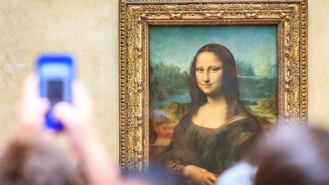 Aufnahme des Ölgemäldes "Mona Lisa" von Leonardo da Vinci im Louvre Museum in Paris – im Bildvordergrund sind Besucher zu sehen.