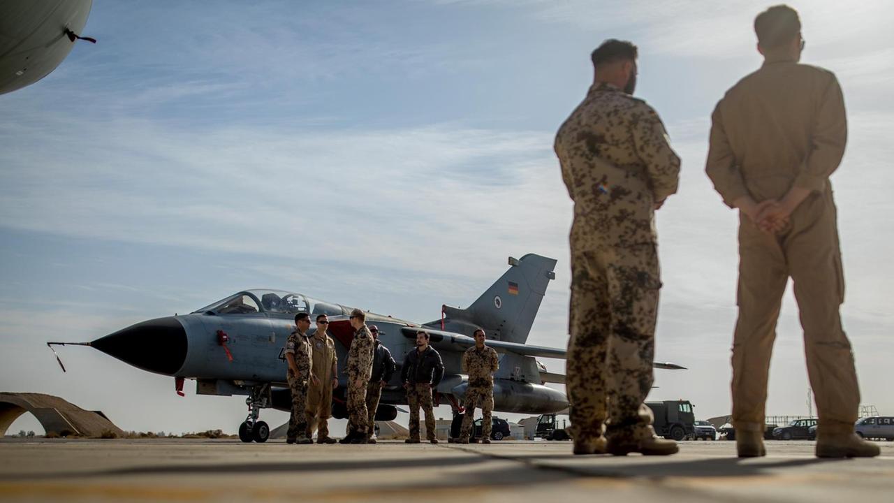 Der Kampfjet steht auf einem Flugplatz, daneben mehrere Soldaten. Im Vordergrund zwei weitere, die mit dem Rücken zur niedrig positionierten Kamera stehen. 