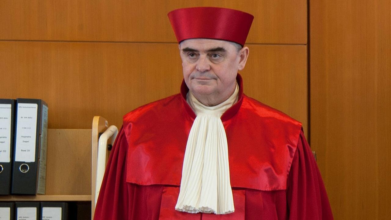 Müller in roter Robe und Mütze steht vor einer Wand mit einem Regal voller Ordner.