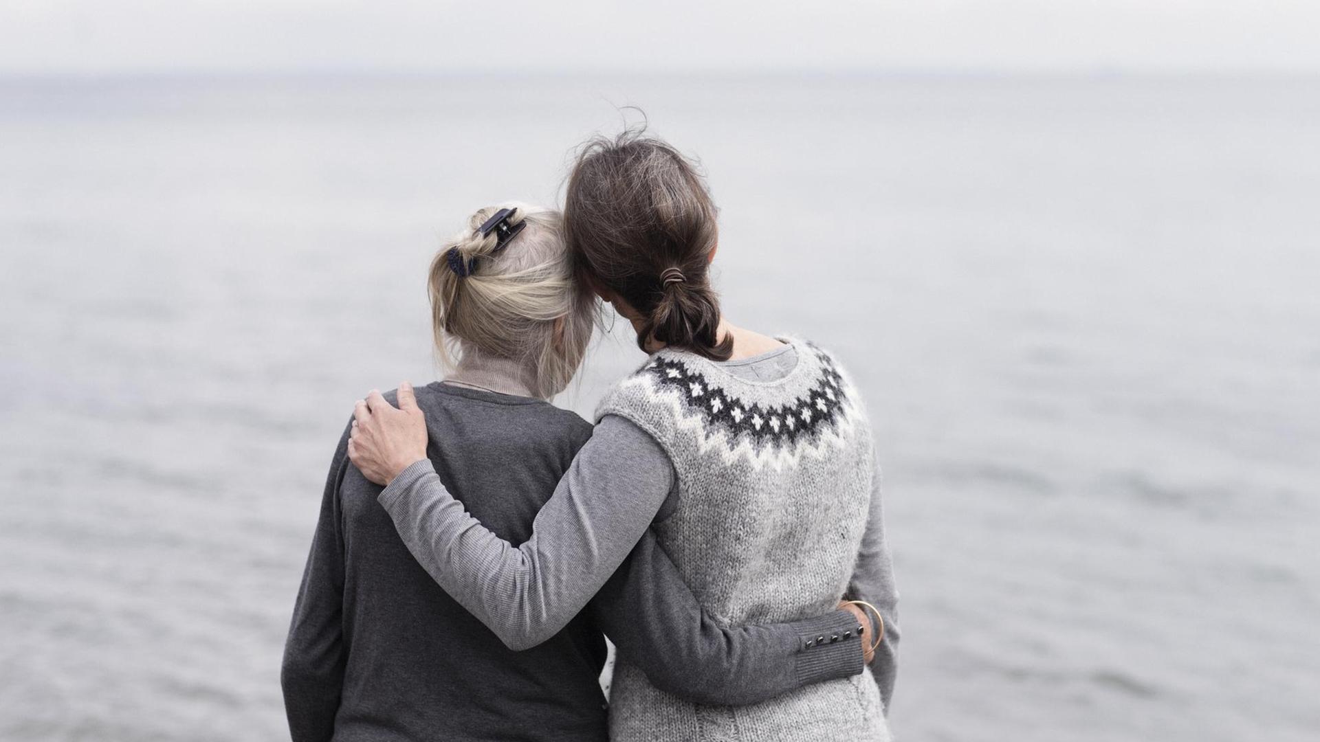 Zwei erwachsene Frauen, Mutter und Tochter stehen Arm in Arm am Meer.