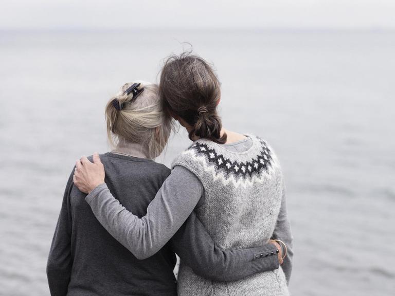 Zwei erwachsene Frauen, Mutter und Tochter stehen Arm in Arm am Meer.