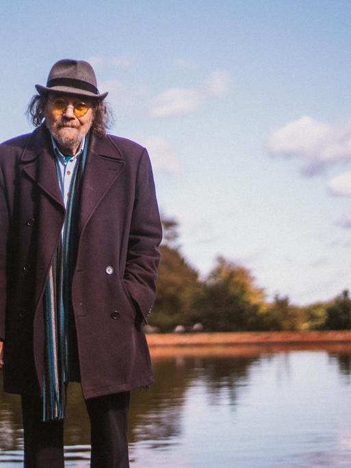 Bill Fay steht mit gelb getönter Sonnenbrille, Hut und Mantel vor einem Teich.