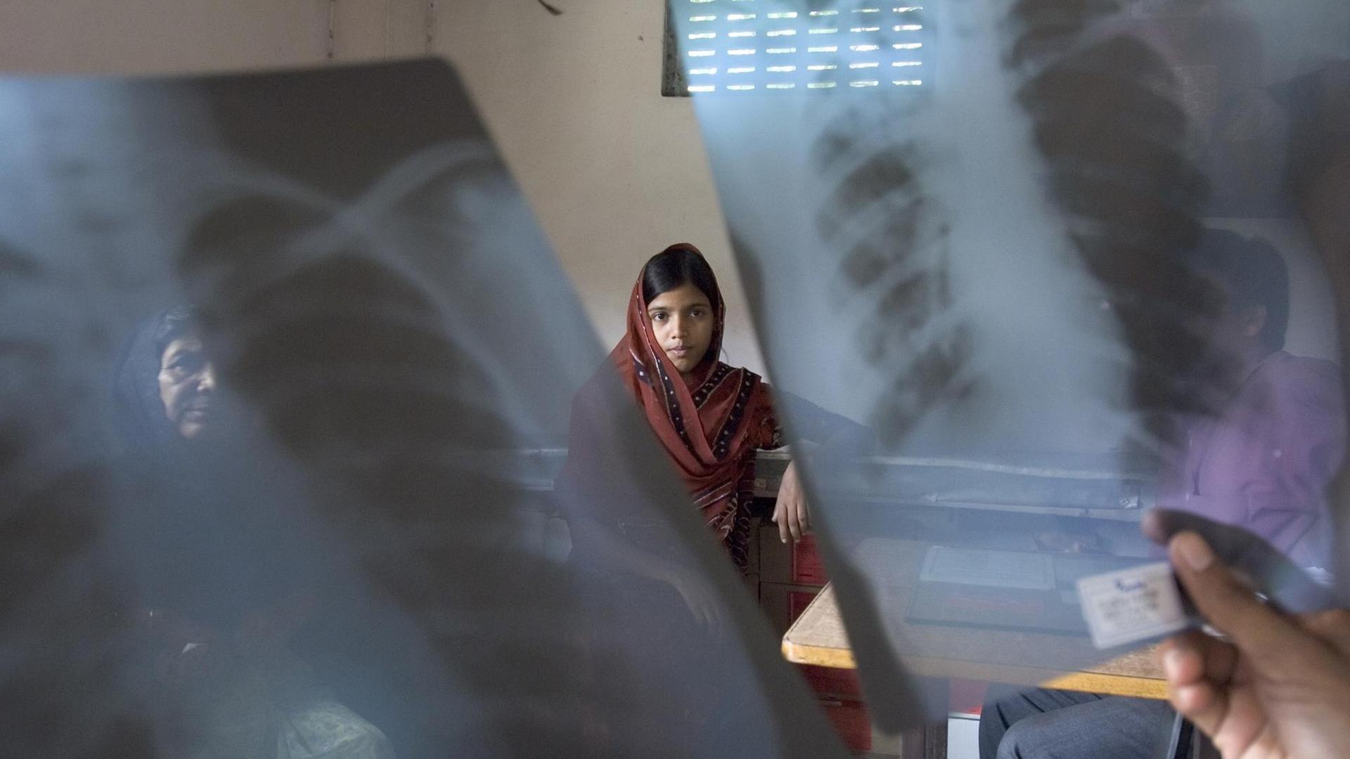 Ein Mädchen sitzt an einem Tisch und blickt in die Kamera. Im Vordergrund hält eine Person zwei Röntgenaufnahmen einer Lunge in die Luft.