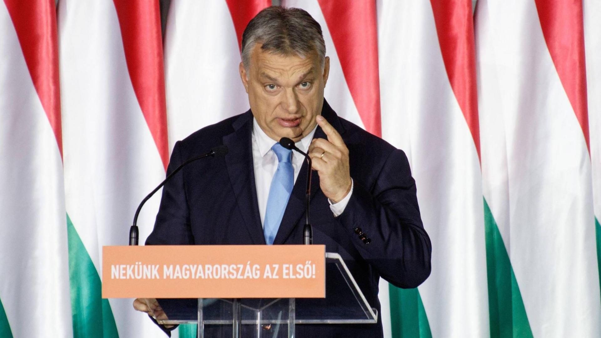 Ungarns Ministerpräsident Viktor Orbán steht bei einer Rede an einem Pult und hebt die linke Hand