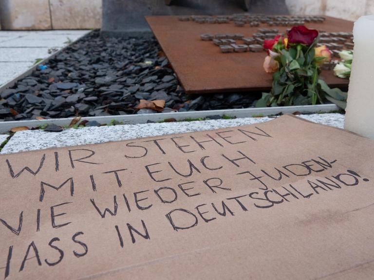 Ein Pappkarton mit der Aufschrift «Wir stehen mit euch nie wieder Judenhass in Deutschland!», Blumen und Kerzen sind vor einer Synagoge abgelegt.