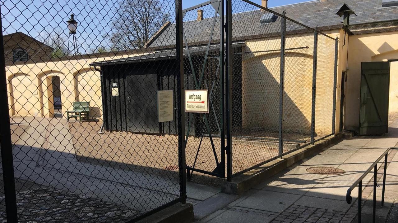Das Bild zeigt einen Hof, der hinten von einer Mauer und vorne durch einen Zaun umgrenzt ist. Auf einem Tor im Zaun ist auf einem Schild "Eingang" zu lesen.