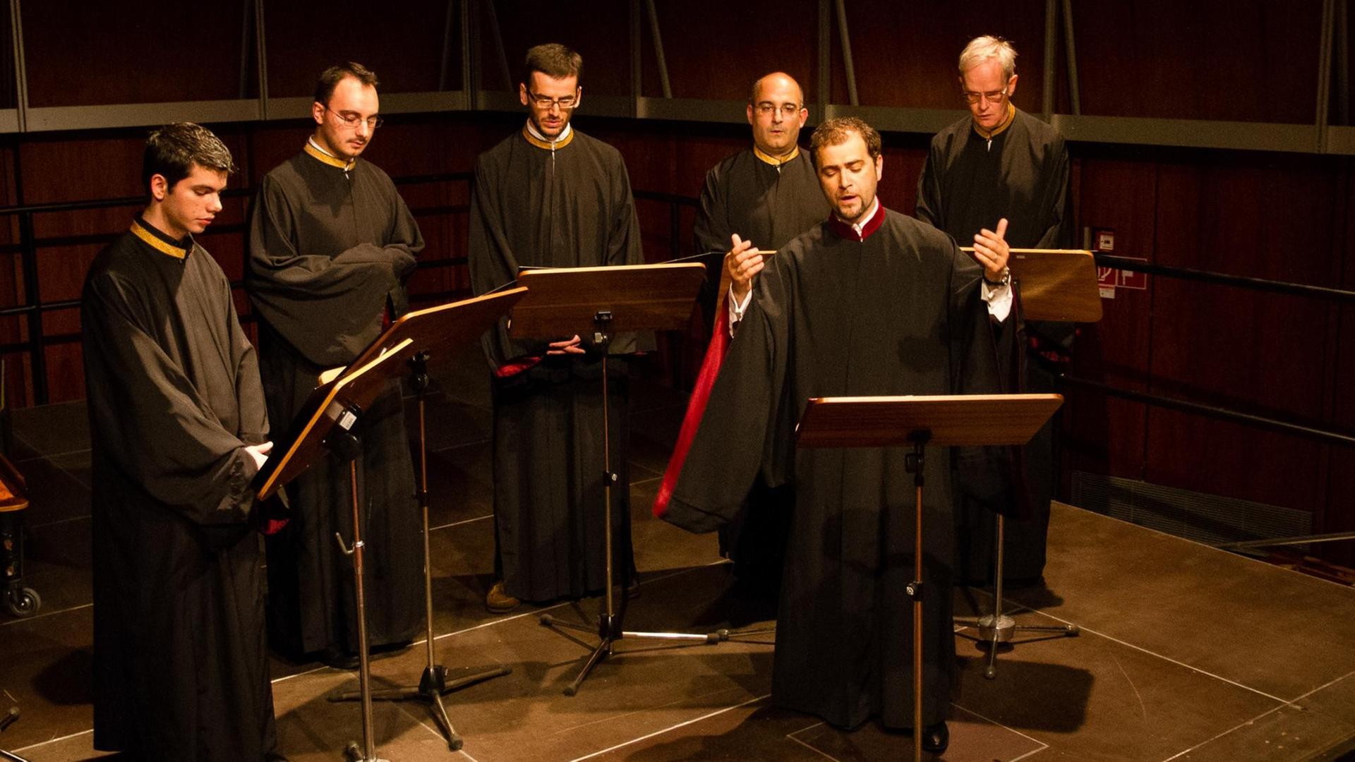 Sechs Männer in Roben stehen vor Notenständern auf einer Bühne.