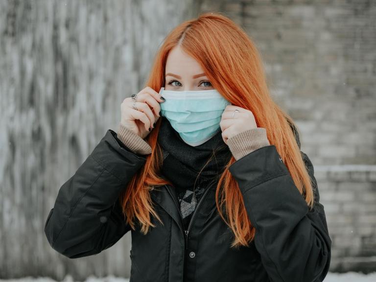 Eine rothaarige Frau in Winterjacke trägt eine Schutzmaske und schaut skeptisch.